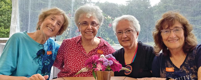 Four smiling women sit at a table with a wooded area visible behind them